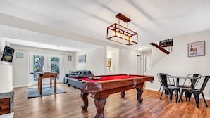 Una habitación amplia con mesa de billar, zona de comedor con sillas y una escalera que sube a otro nivel.