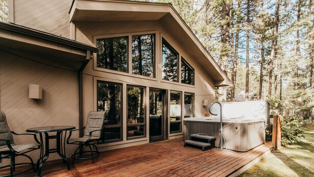 A wooden deck with outdoor seating, a hot tub, and large windows overlooking a forested area. - A Lovely home, Pet Friendly, Hot Tub & National Forest (Black Butte Ranch)