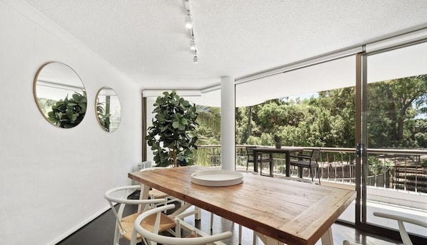 Un espace de salle à manger moderne avec une table en bois, des chaises blanches, de grandes fenêtres et un balcon agrémenté de mobilier d’extérieur.