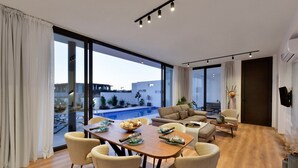 A modern living room with a dining area, large windows, and a view of a swimming pool. - Protaras Almaria Villa 16 (Protaras)