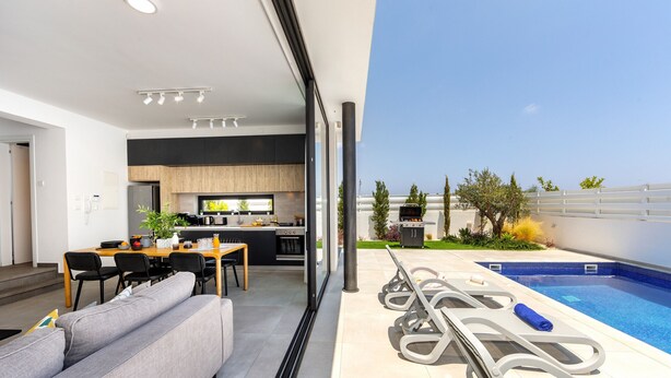 A modern kitchen with a dining area, a poolside lounge, and a view of the pool. - Protaras Almaria Villa 16 (Protaras)