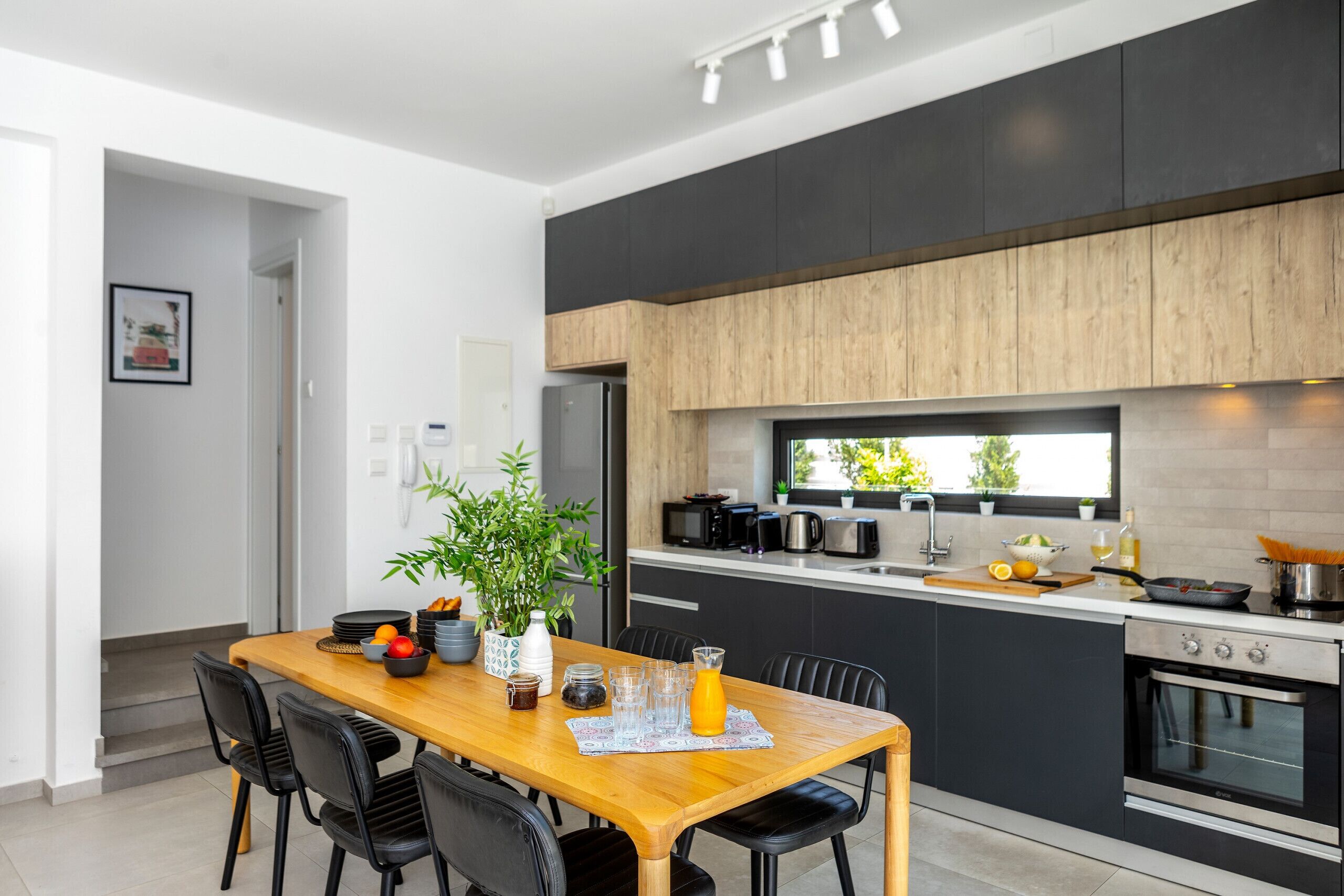 Una cocina moderna con armarios oscuros, encimera de madera y horno integrado. Hay un comedor con mesa de madera y sillas negras.