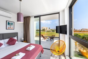A hotel room with a bed, a TV, a balcony with a chair, and a view of buildings. - Protaras Almaria Villa 16 (Protaras)