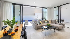 A modern living room with a sofa, coffee table, dining table, and chairs. There is a large window with curtains, a blue pool visible outside, and a plant on the table. - Protaras Almaria Villa 16 (Protaras)