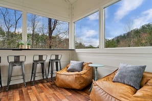 Una habitación soleada con ventanas amplias, piso de madera, un sillón de cuero tipo orejero y taburetes de metal.