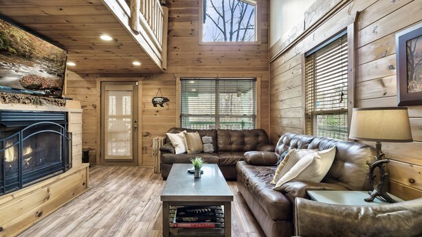 A cozy cabin interior with a fireplace, leather sofas, a coffee table, and a large window with blinds. - ⛰Mtn Views, 🔥Fire Pit, Hot Tub, 🕹Game/Movie Room (Sevierville)