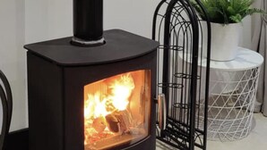 A black wood-burning stove with a visible fire, a black metal chair, a white round table, and a potted plant. - Luxurious Studio Apartment in Fahan, Co Donegal (Fahan)