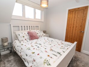 A bedroom with a white bed, floral bedding, a wooden door, and a window with a light fixture.