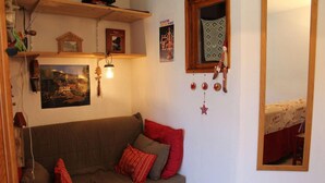 Un salón acogedor con un sofá marrón, cojines rojos y grises, una estantería de madera con libros y una maqueta de una casa, un cuadro enmarcado y una ventana con cortina estampada.