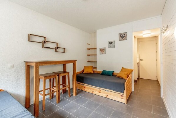 A compact room with a bed, a small wooden desk, and a chair. There are two wall-mounted shelves and a door leading to another room.