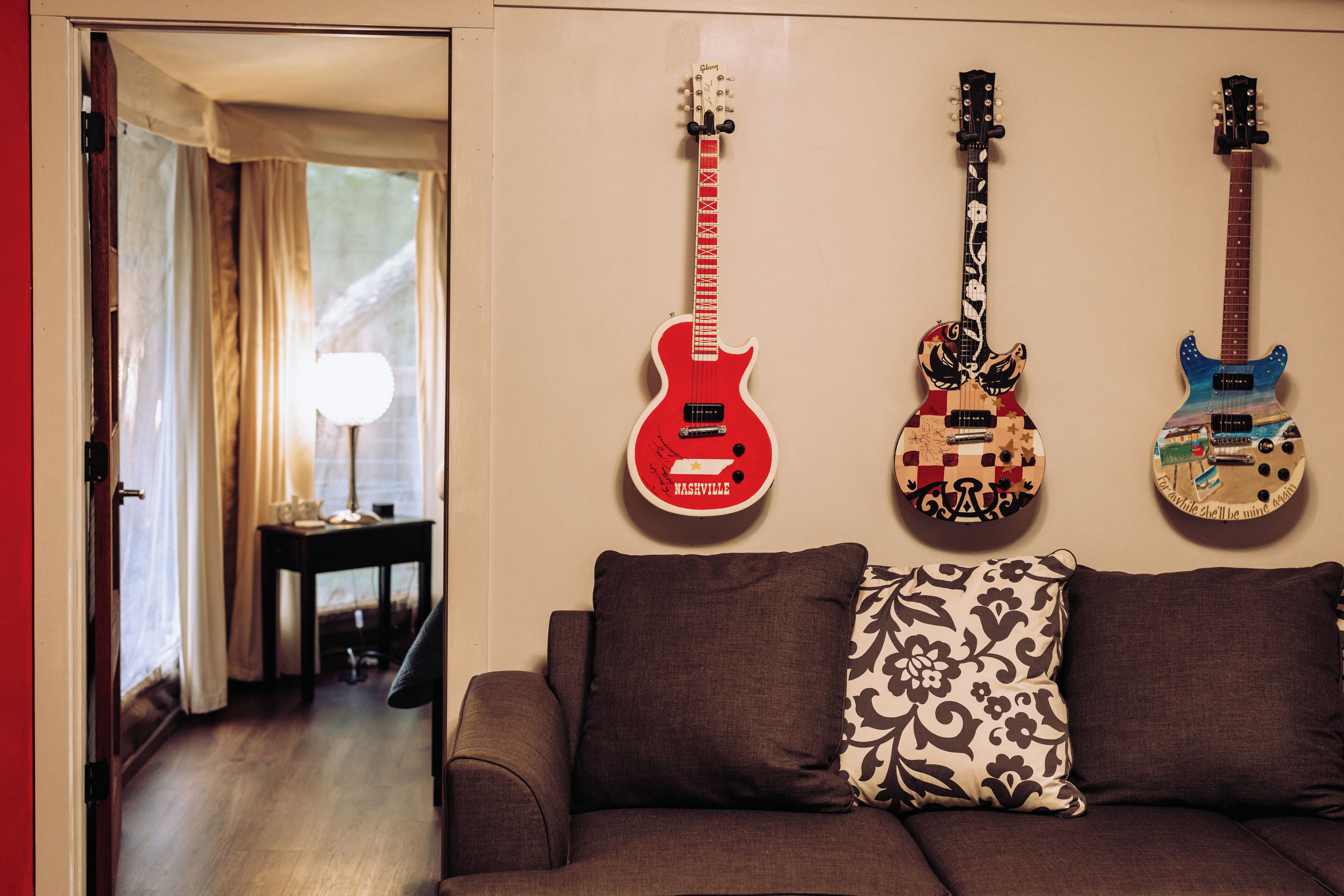 A room with a sofa, three guitars on the wall, and a lamp on a side table.