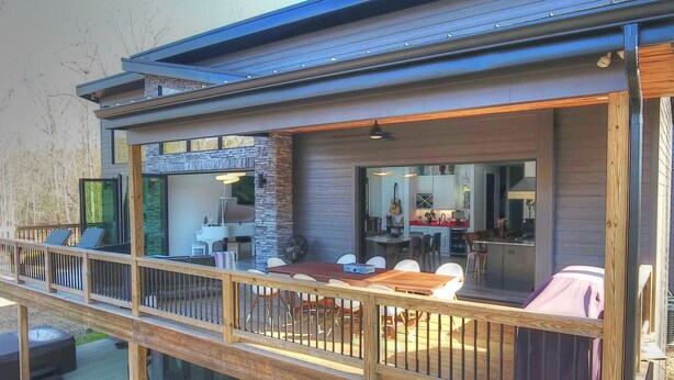 Una terraza moderna al aire libre con zona de comedor, una cocina visible a través de un ventanal y baranda de madera.