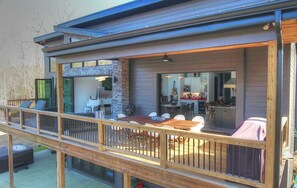 A modern outdoor deck with a dining area, a kitchen visible through an open door, and a wooden railing.