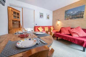 Tunnelmallinen olohuone, jossa on punainen sohva, puinen ruokapöytä kahdelle ja lasiovilla varustettu puinen kaappi.