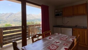 Zona pranzo con tavolo ricoperto da tovaglia fantasia, sedie in legno, vista sulle montagne tramite un balcone e cucina con mobili in legno e forno a microonde.
