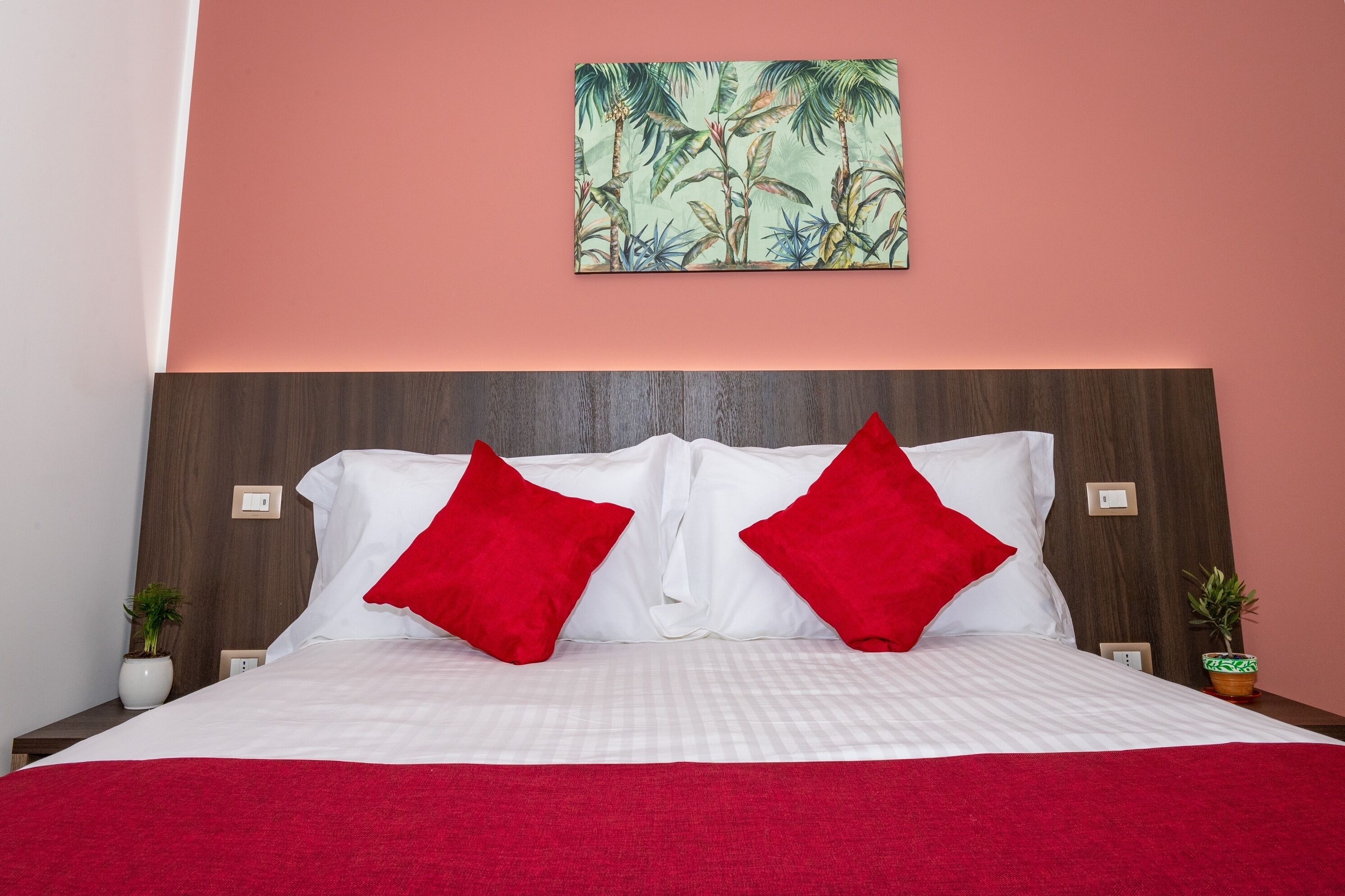 A neatly made bed with white linens and two red pillows, a wooden headboard, and a tropical-themed painting on the wall.