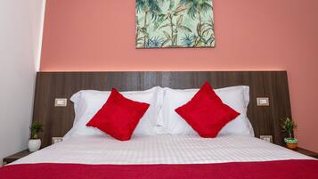 A neatly made bed with white linens and two red pillows, a wooden headboard, and a tropical-themed painting on the wall.