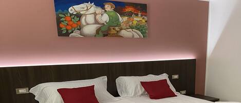 Una cama doble con sábanas blancas y almohadas rojas, una mesita de noche y un cuadro colgado en la pared.