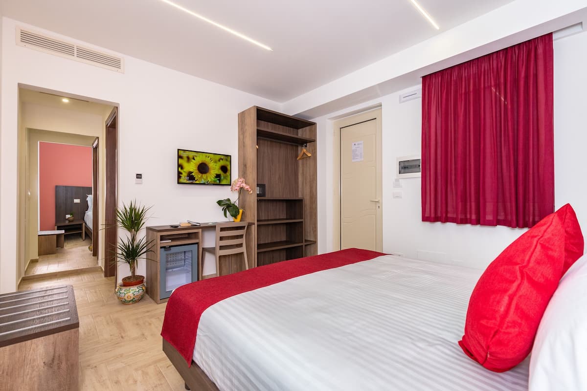 Ein Hotelzimmer mit einem Bett, einem Schreibtisch, einem Kleiderschrank, einer Zimmerpflanze und einem roten Vorhang.