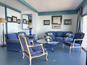A room with a blue sofa set, matching armchairs, a round glass coffee table, and framed artwork on the walls.