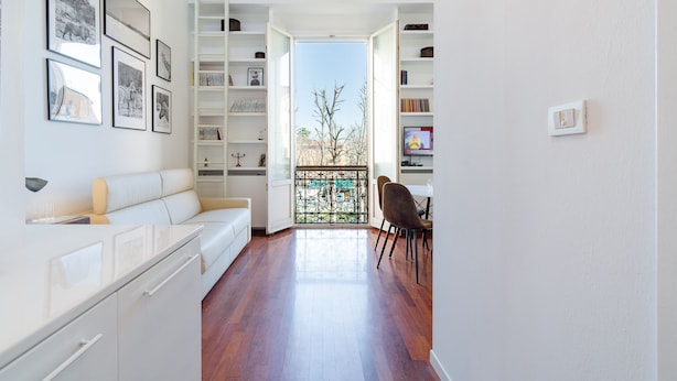 A modern, open-plan living space with a white sofa, dining area, and a view through an open door to a balcony.