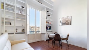 A modern living room with a white sofa, a dining area with a small table and chairs, and built-in shelving with books and decorative items.