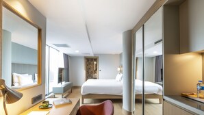 Une chambre d’hôtel moderne équipée d’un lit, d’un bureau avec une lampe, d’une chaise et d’un grand miroir.