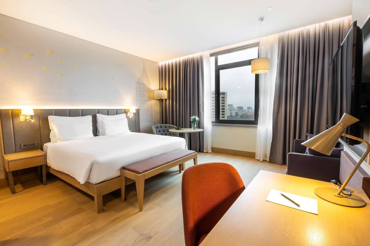 A hotel room with a bed, bedside tables, a desk with a lamp, a chair, and a view of the city through large windows.