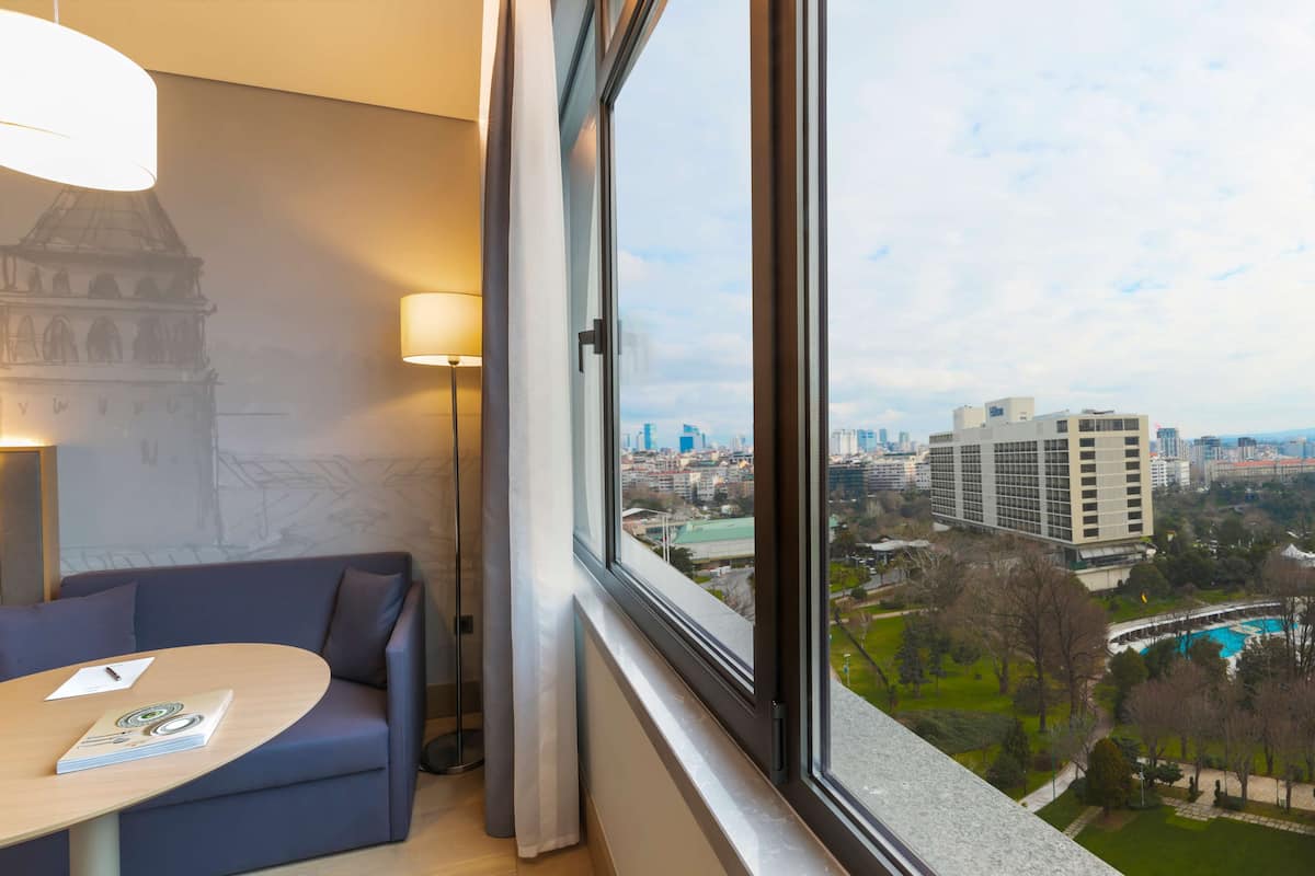 A modern hotel room with a large window offering a city view, a comfortable sofa, a small table, and a lamp.