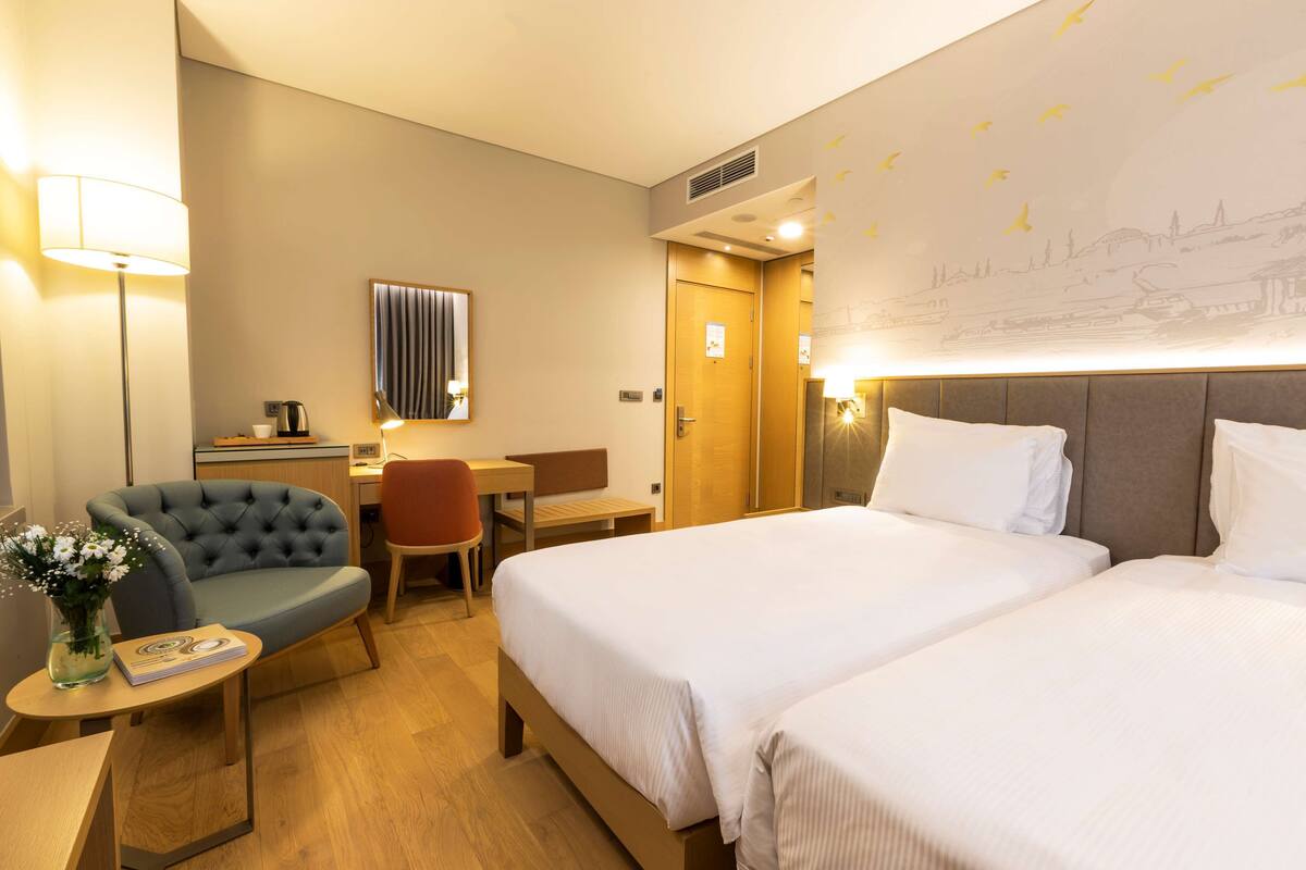 A hotel room with two beds, a desk, a chair, and a lamp.