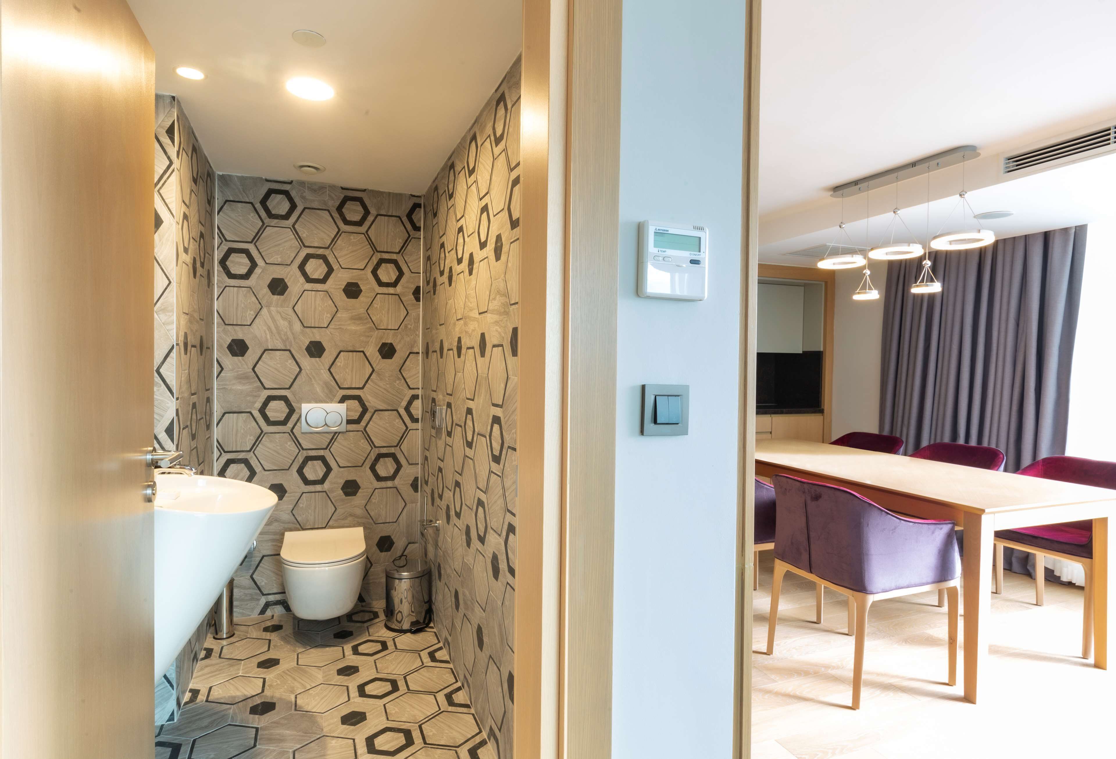 Un baño moderno con un patrón de baldosas hexagonales, un lavabo blanco y un inodoro. Se puede ver un comedor con una mesa de madera y sillas moradas a través de una puerta abierta.
