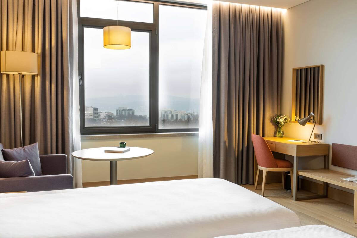 A hotel room with a bed, a desk with a chair, a small table, and a view of a cityscape through a large window.