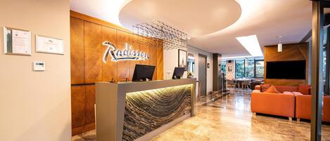 A reception area with a marble floor, a reception desk with a wood finish, and a modern chandelier. There is a seating area with orange sofas and a flat-screen TV mounted on the wall.
