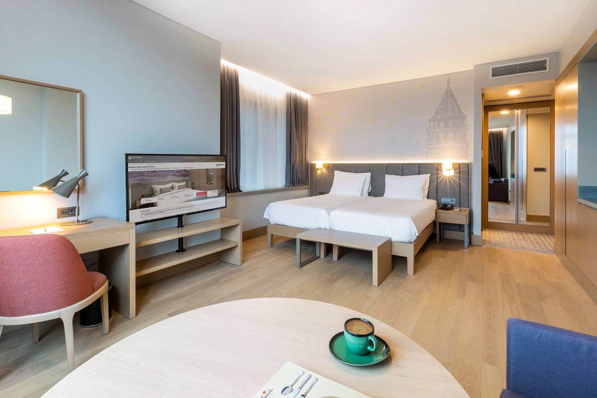 A hotel room with a bed, desk, chair, and a table with a cup.