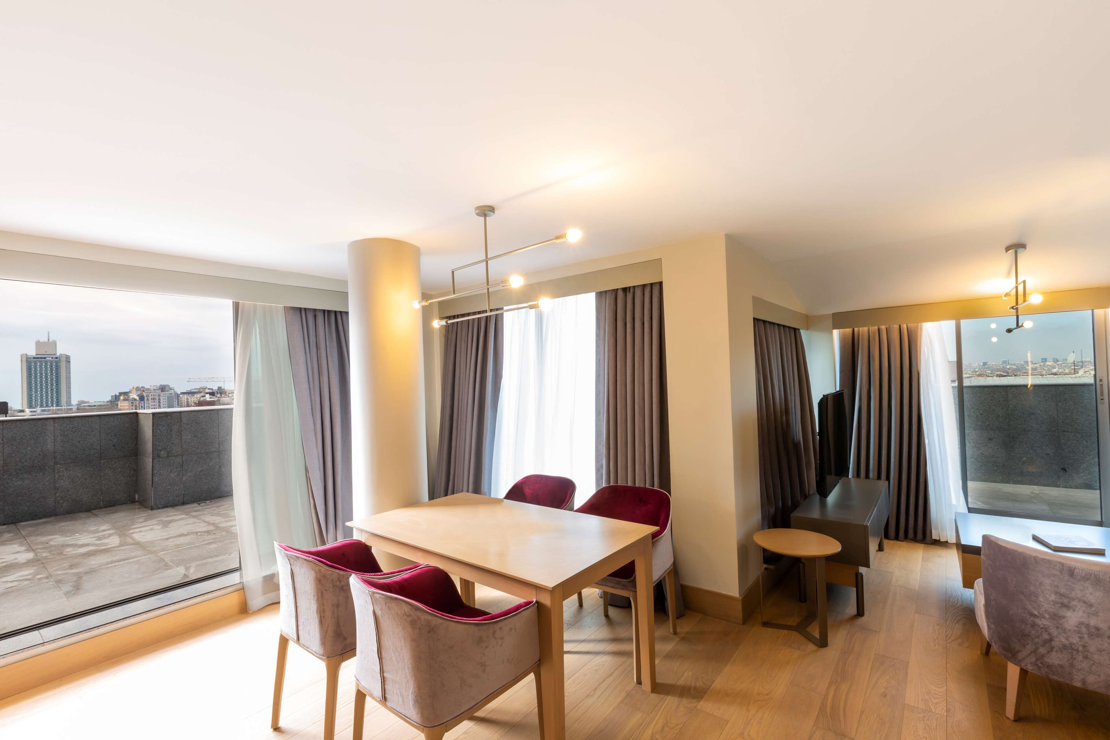 Uma sala de jantar moderna com mesa de madeira e cadeiras vermelhas, janelas amplas com cortinas e vista para a cidade.