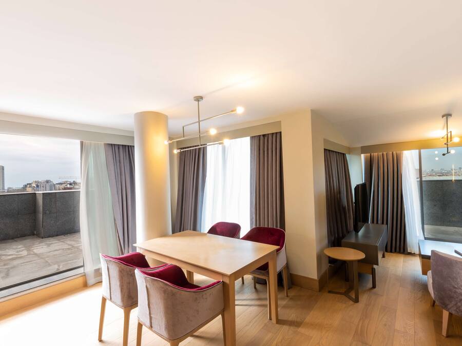 Uma sala de jantar moderna com mesa de madeira e cadeiras vermelhas, janelas amplas com cortinas e vista para a cidade.