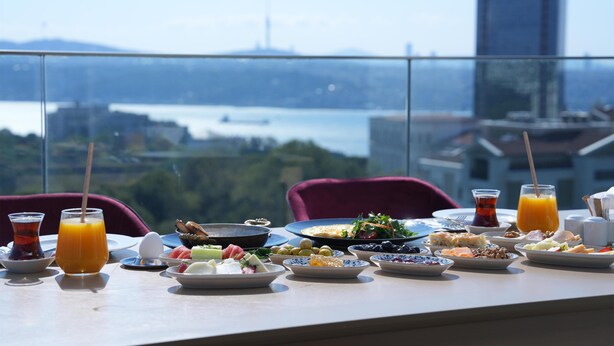 Une table dressée avec des aliments pour le petit-déjeuner et un verre de jus d’orange, donnant sur un paysage urbain avec un plan d’eau et un grand immeuble.