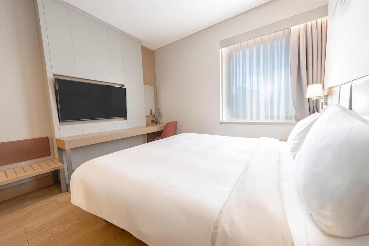 A hotel room with a large bed, a TV mounted on the wall, a bedside table, a window with curtains, and a bench by the window.