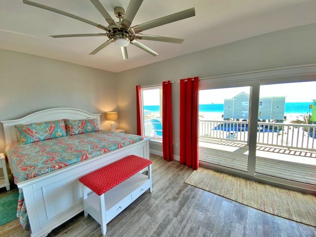 Une chambre à coucher avec un lit, un banc, un ventilateur de plafond et une vue sur l’océan grâce à de grandes fenêtres.