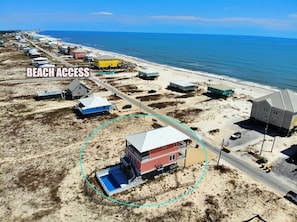 Luchtbeeld van een strandgebied met meerdere gebouwen, waaronder een opvallend rood gebouw met een blauw zwembad op de voorgrond, en een weg die naar het strand leidt.