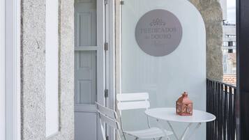 Un balcón con una silla plegable blanca, una mesita redonda pequeña y un farol. Se ve una puerta al fondo y un cartel en la pared.