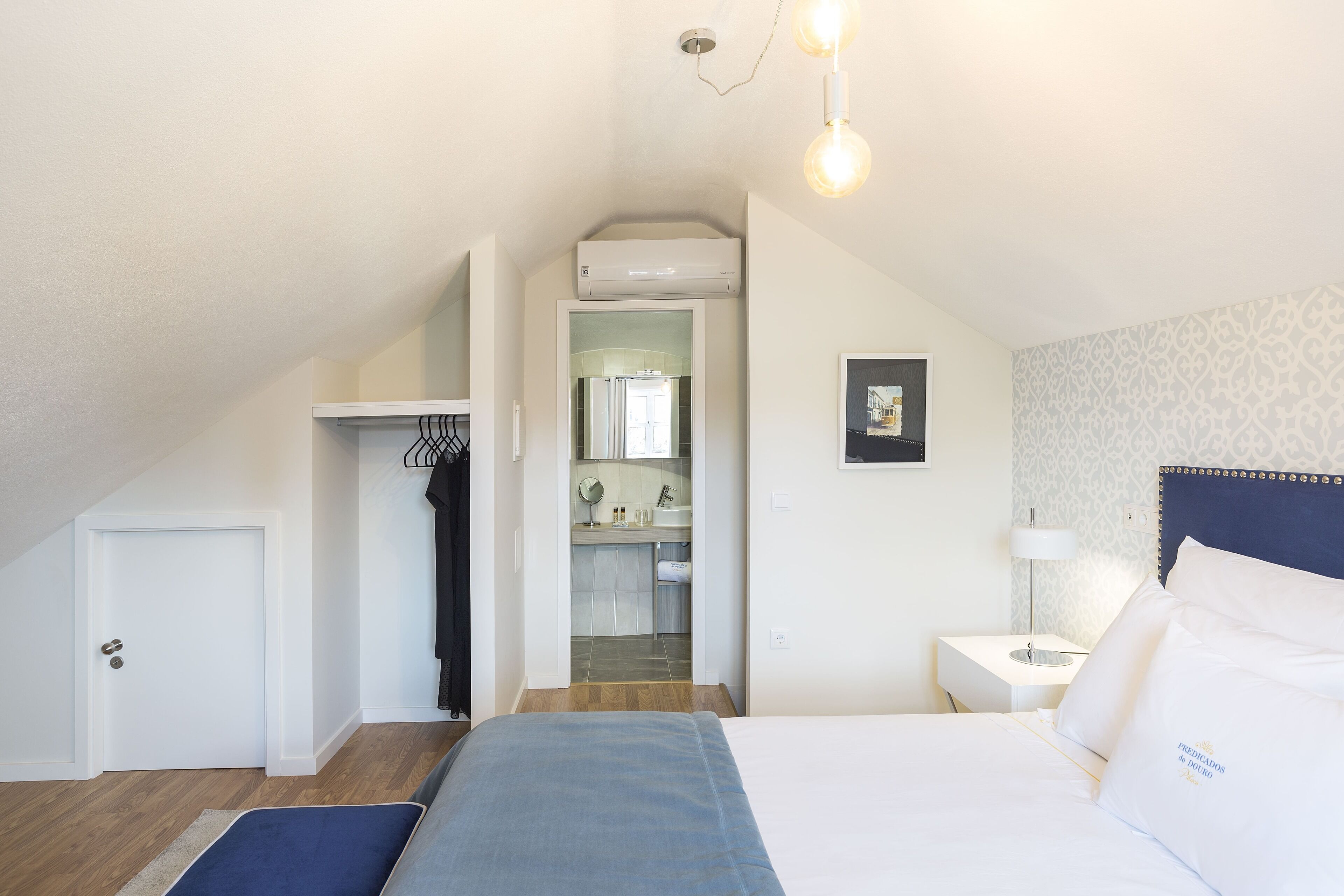 A bedroom with a bed, a nightstand, a lamp, a wardrobe, and a bathroom visible through an open door.