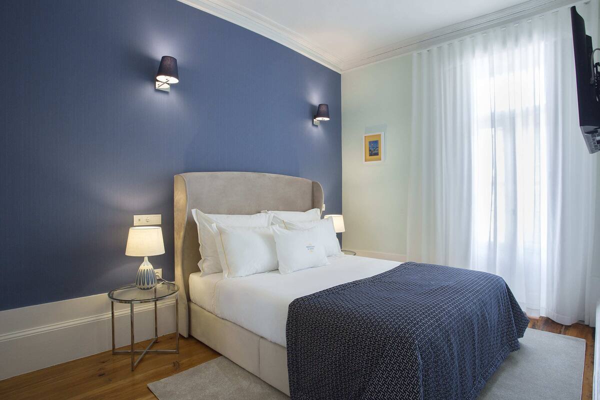 A bedroom with a bed, two bedside lamps, a small table, a rug, and a window with curtains.