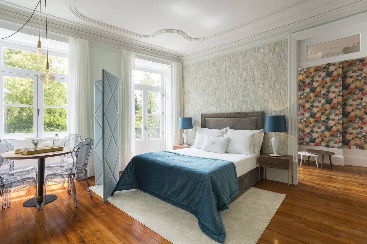 A bedroom with a large bed, a desk, and a chair. There is a window with curtains, a door, and a wall with a floral pattern.