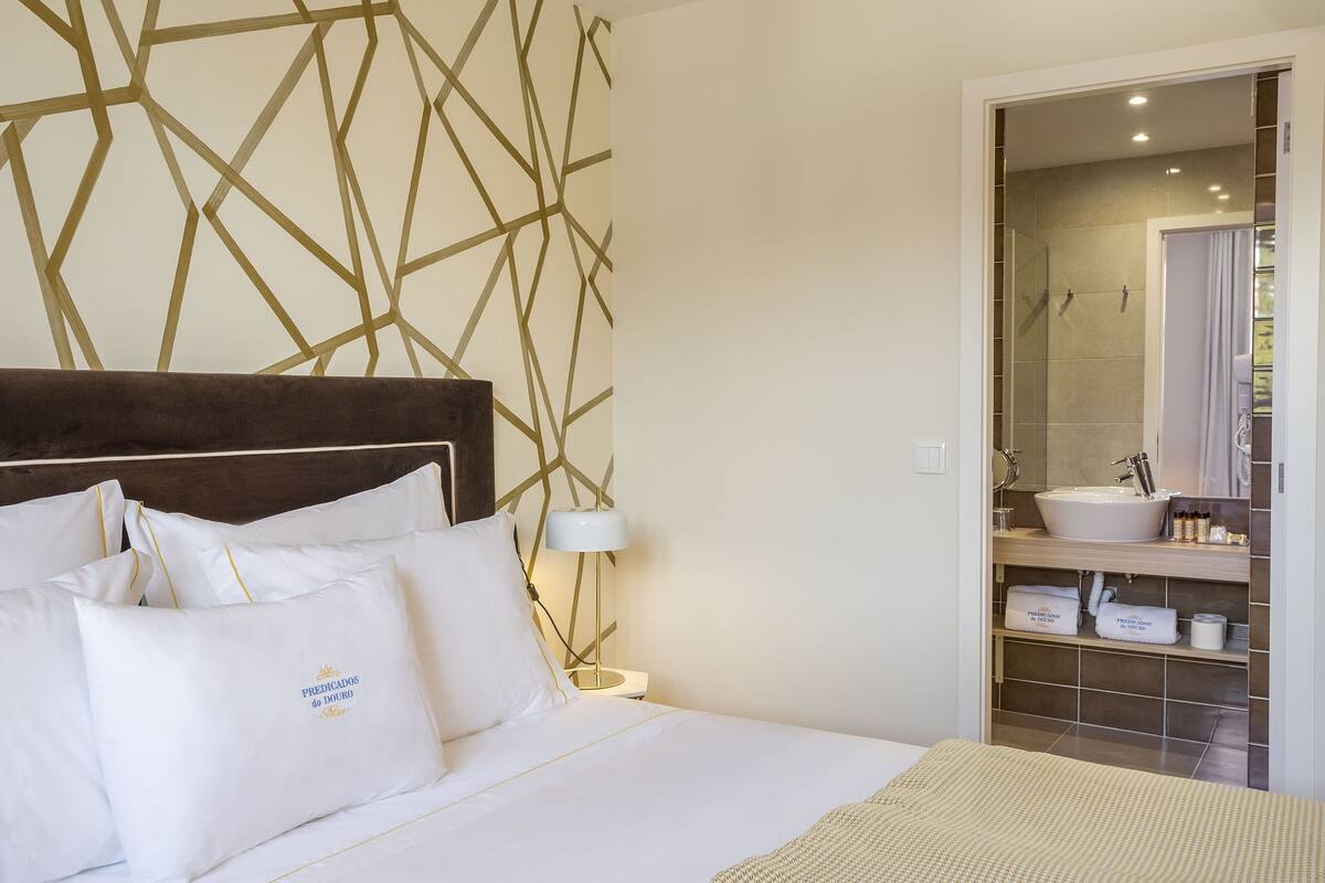 A hotel room with a neatly made bed, a bedside lamp, a bathroom with a sink and mirror, and a geometric wall design.