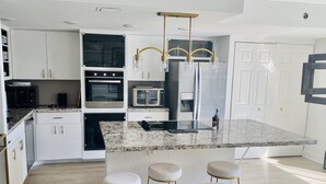 A modern kitchen with white cabinetry, a granite countertop, a built-in oven, a microwave, and a stainless steel refrigerator. There are three bar stools and a marble island. - Lovely two story home on the water (Hallandale Beach)