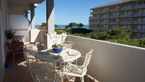 A balcony with a white table and chairs, a potted plant, and a view of a yellow building and the sea.
