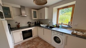A compact kitchen with white cabinets, a built-in oven, a washing machine, and a window with a view of greenery. - Charming Penty Breton, close to the beaches and town of Tregunc - 6 people max. (Trégunc)