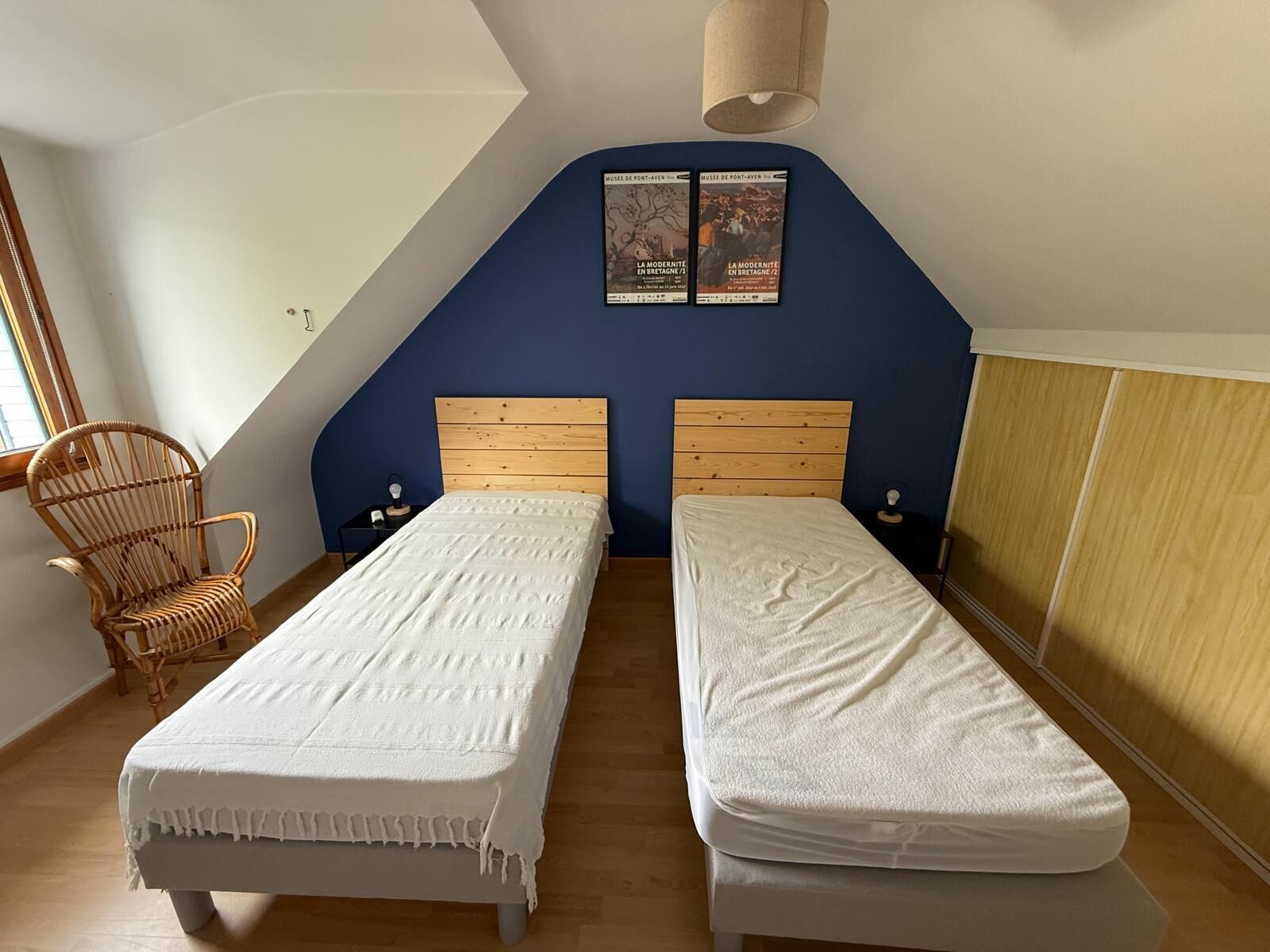 Een slaapkamer met twee bedden, een houten stoel, een lamp en twee posters aan de muur.