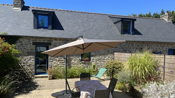 A stone house with a tiled roof, a patio with a table and chairs, and a large umbrella. - Charming Penty Breton, close to the beaches and town of Tregunc - 6 people max. (Trégunc)